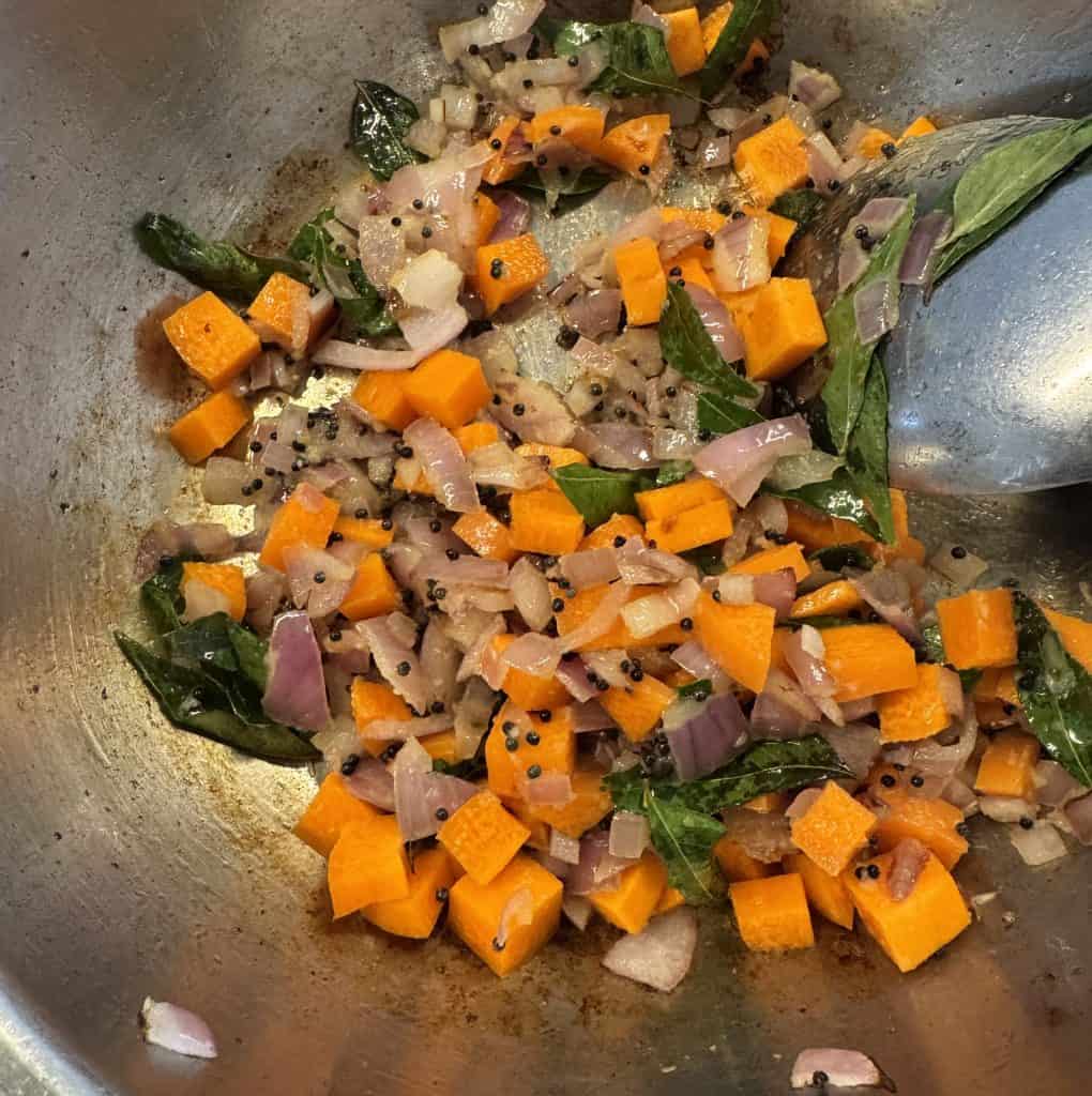 Diced carrots, onions, and curry leaves being sautéed with mustard seeds in a non-stick pan.